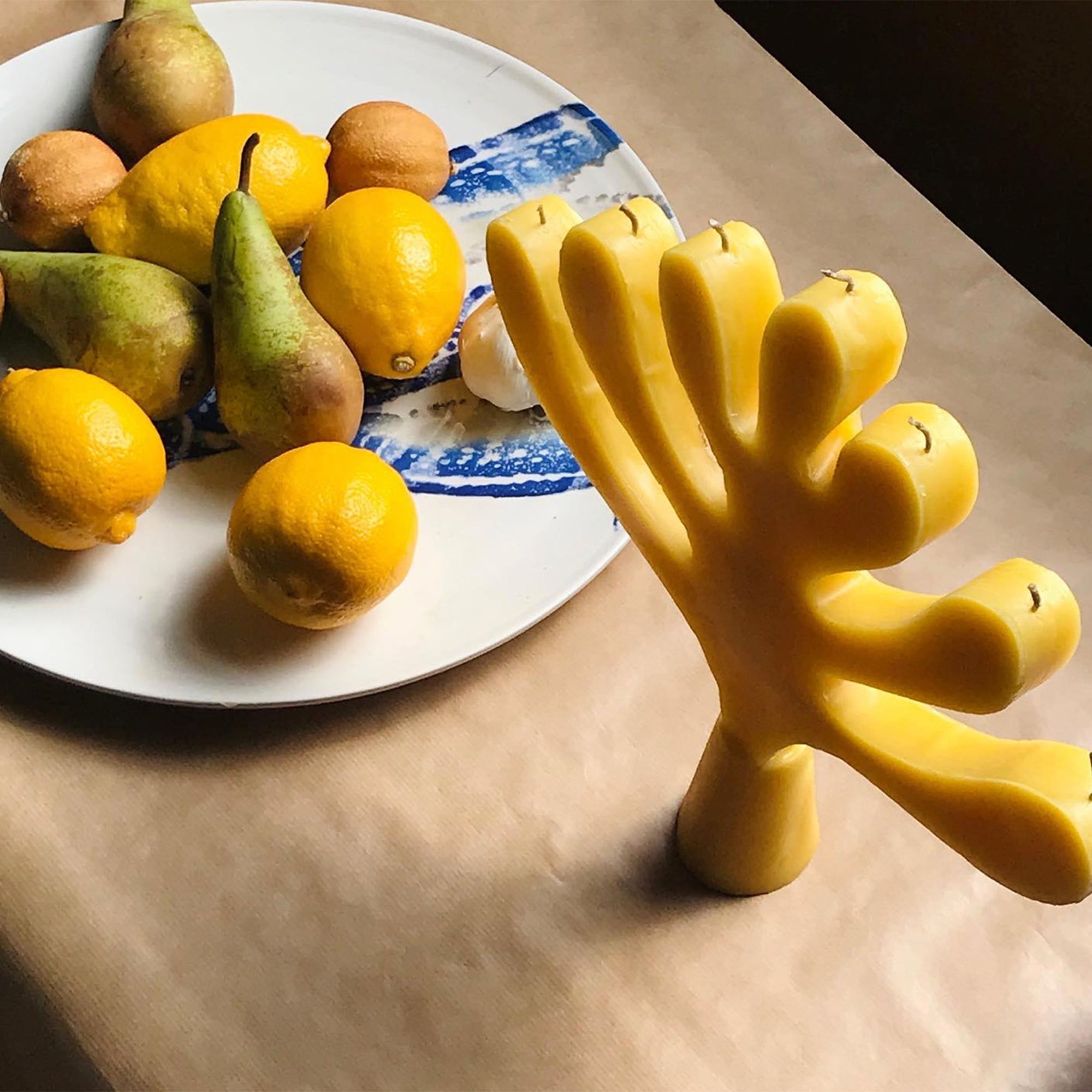 yellow beeswax centerpiece tree of light candle standing on a table next to a white and blue plate filled with pears and lemons.