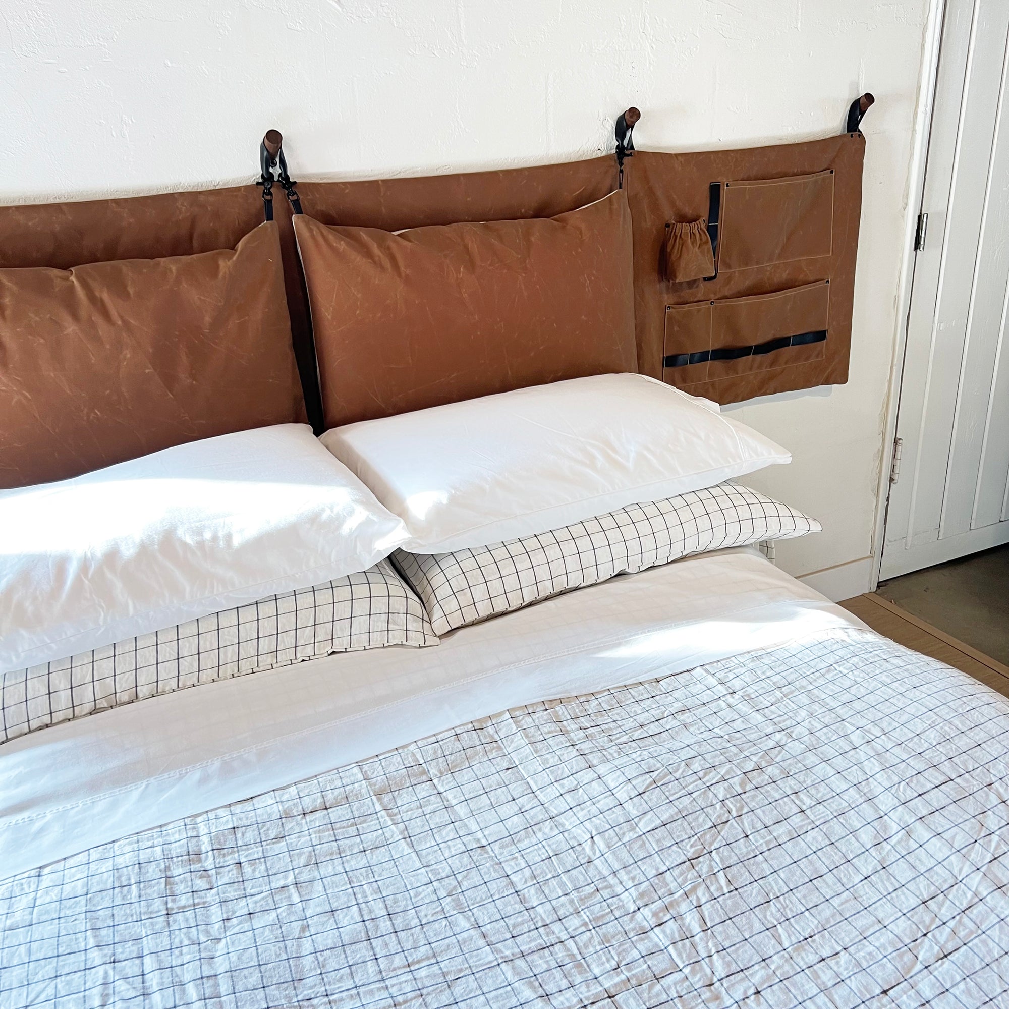 Clean photo of a made bed with linen sheets and a brown waxed cotton canvas headboad hanging on the white wall above the bed. The headboad has black leather and hardware on the side pocket storage panel. 