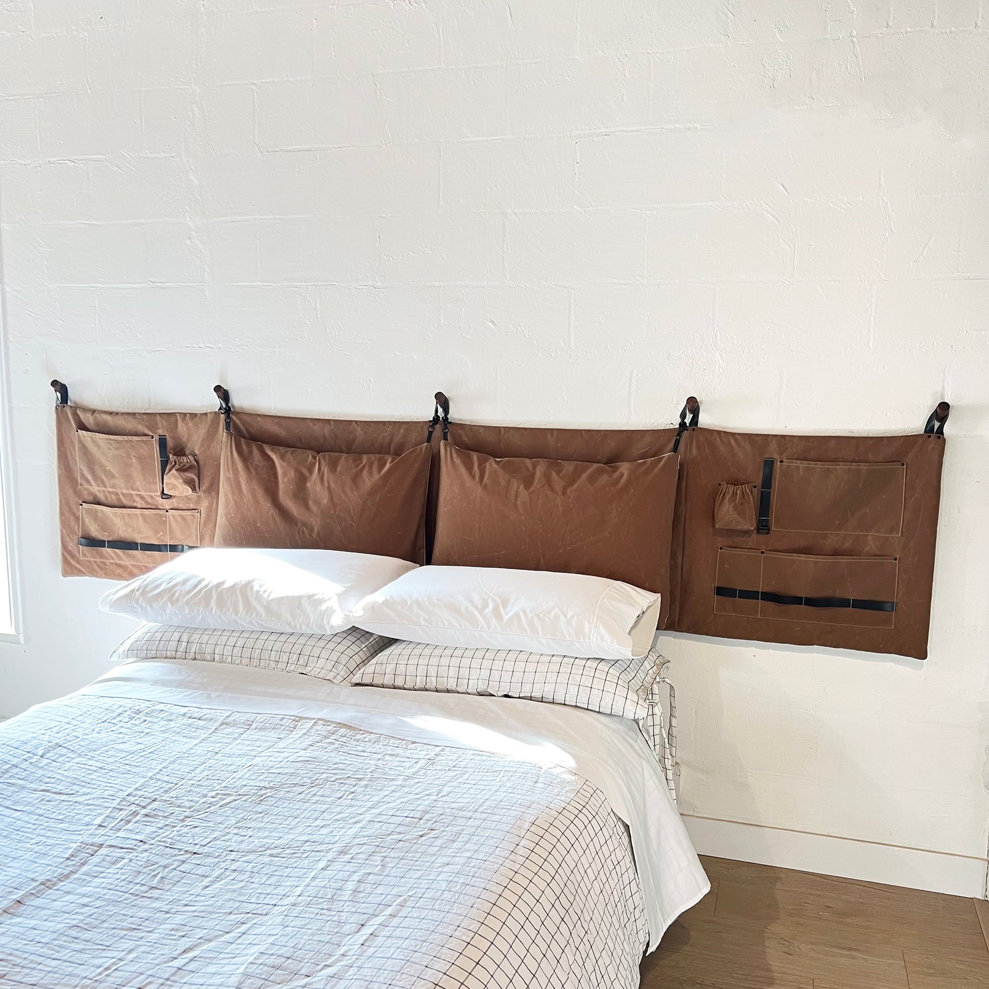 Rugged, camping inspired brown waxed cotton canvas headboard with pockets on the side panels for all your storage needs and matching canvas pillows attached. The headboard is hanging on a white wall over a platform bed with linen bedding.