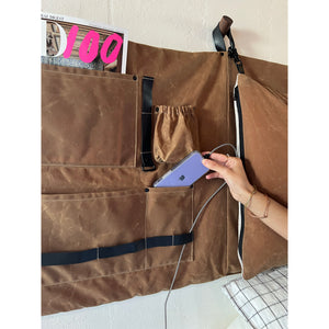 Cotton Waxed Canvas side panel with pockets and black hardware. The top pocket is holding a magazine and there is a hand placing an iphone in another pocket. The iphone charger cord is going through a leather loop. 