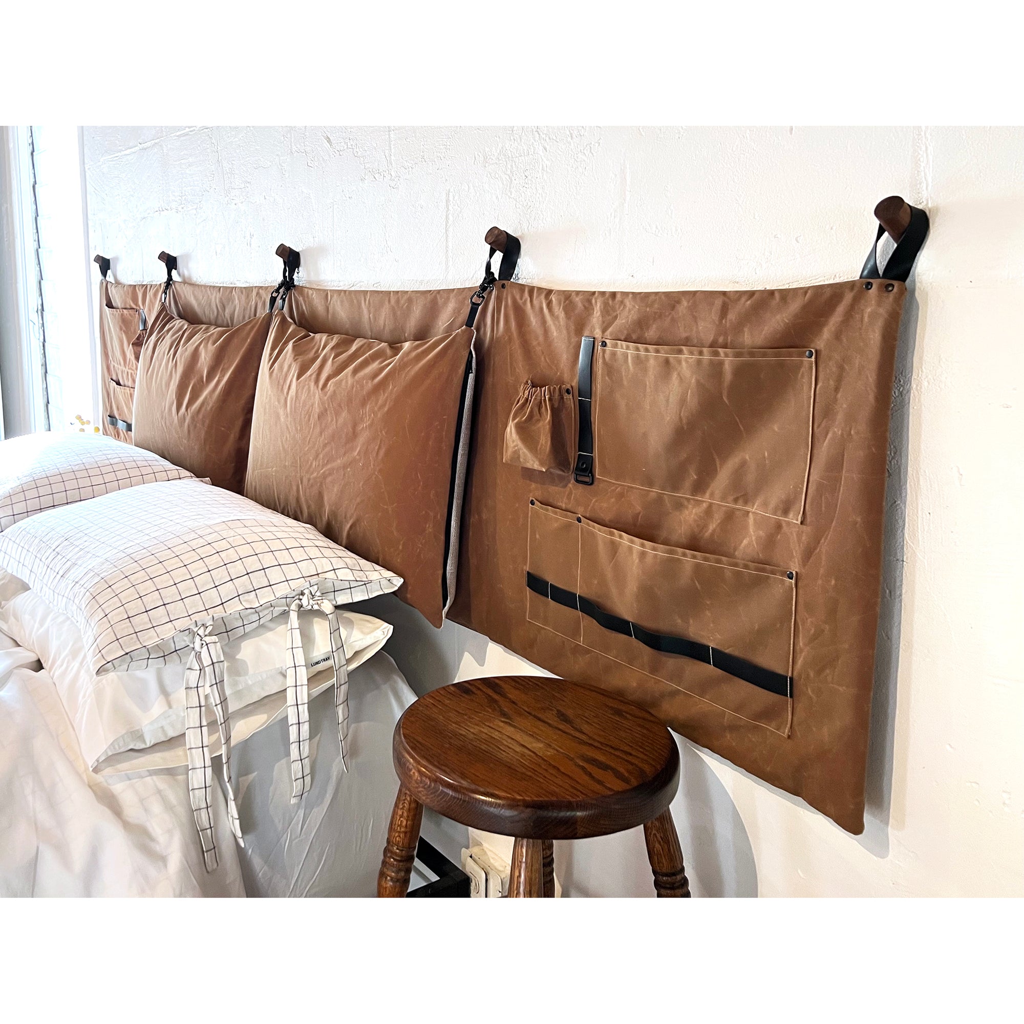 Waxed cotton canvas headboard in camel brown hanging on a white wall over a queen size bed with a wood stool under the pocket side panel. 