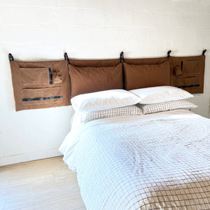 full length scout headboard, camel brown, waxed cotton canvas on a white wall above a made bed. Lots of pillows, pockets and loops for connivence. 