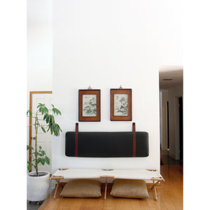 Black leather headboard cushion with straps mounted on a white wall with art above and a cot below. 
