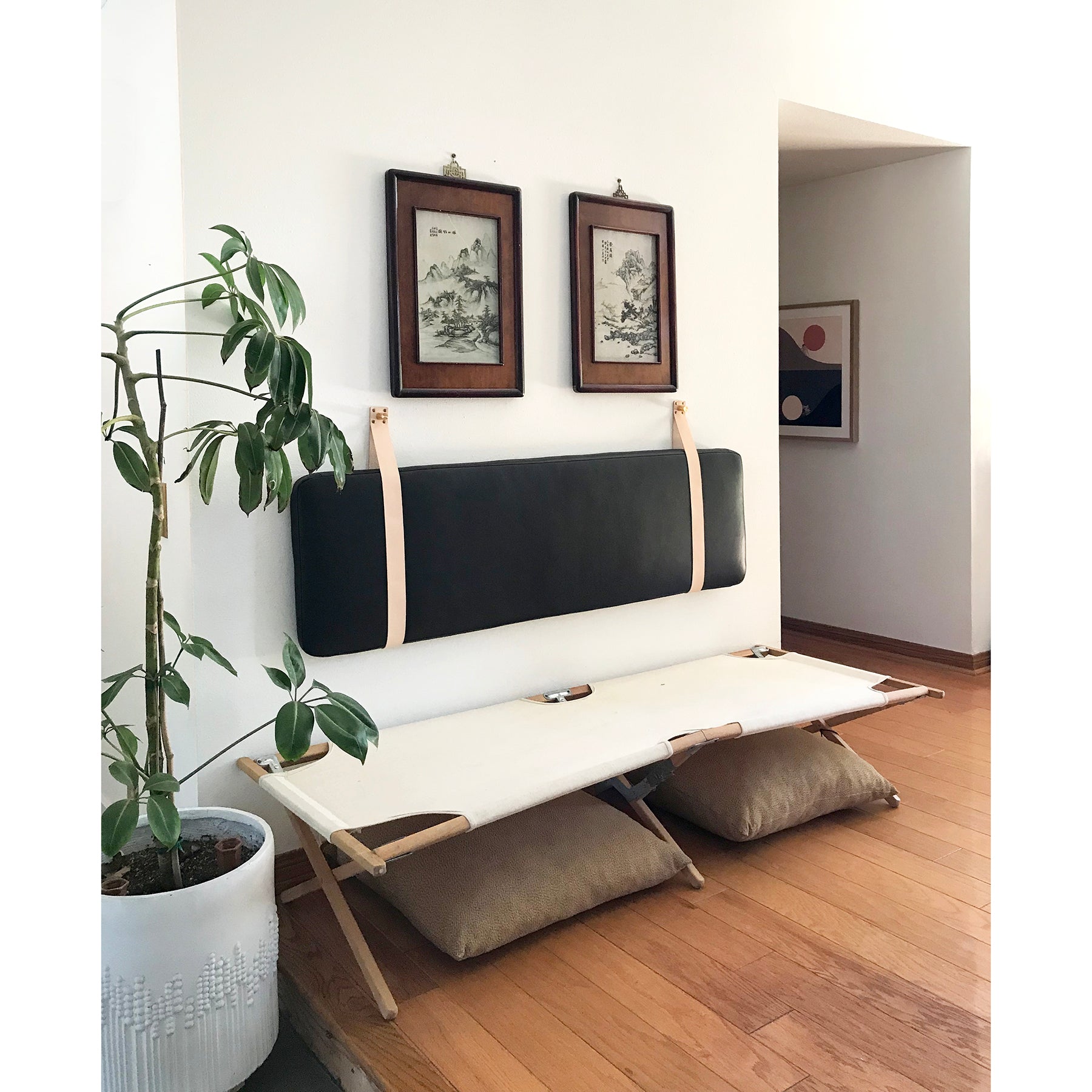 Three quarter view of styled entryway with plant, art and Black leather headboard cushion used as a backrest mounted on a white wall with natural veg-tan leather straps above a cot.