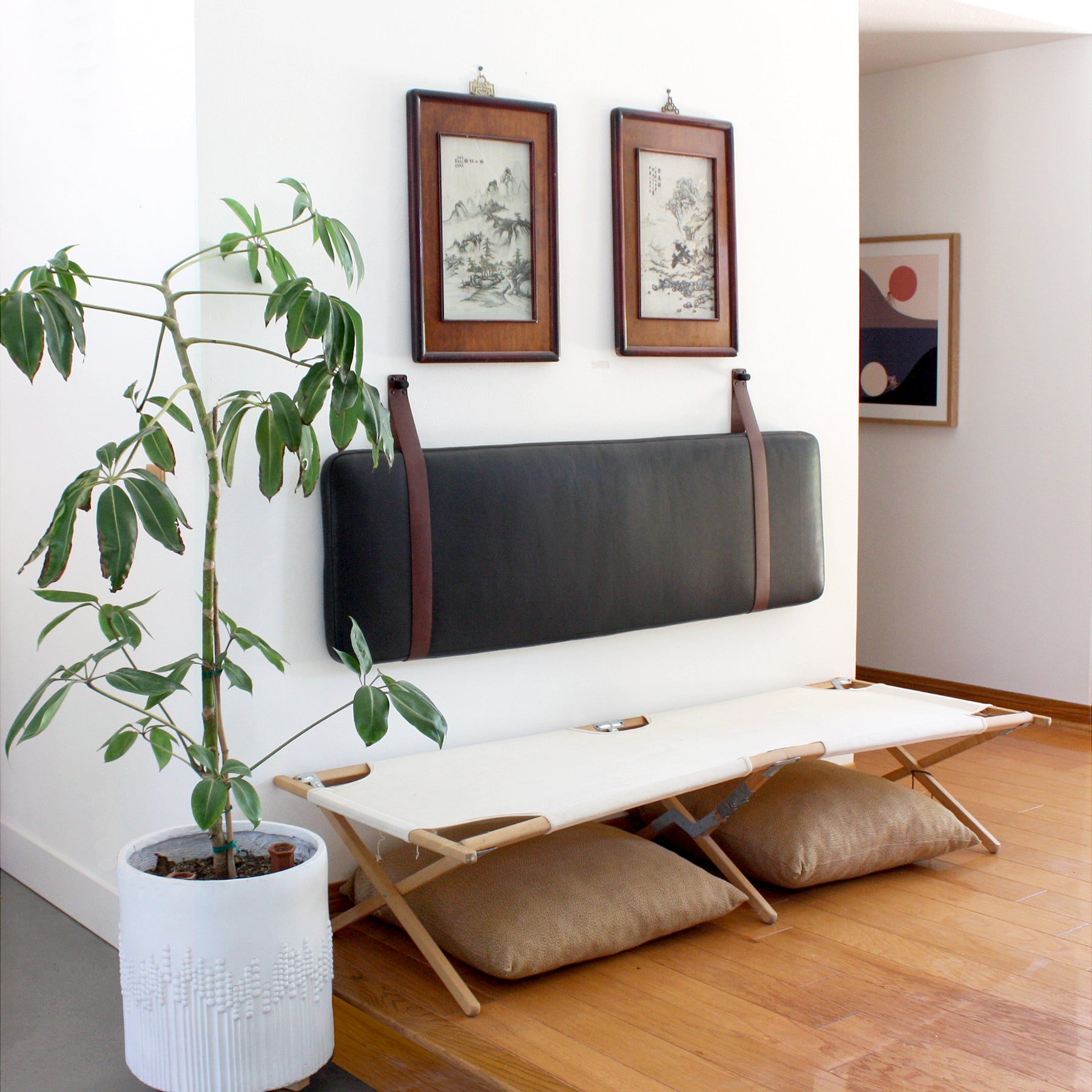Three quarter view of Black Leather Headboard Cushion with Straps mounted on a white wall above a cot in an entryway. Art, pillows and a plant are also in view. 