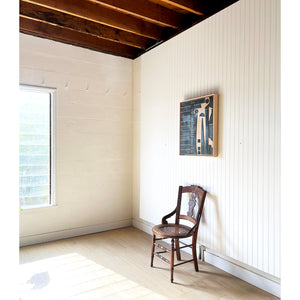 The corner of a room with white walls and wood ceiling. There is a window on the left, a chair on the right, with a work of abstract art by Angie Johnson above the chair