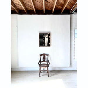 A black leather and wood abstract piece of art hangs on a white wall above a wood chair. Light wood flooring, dark wood ceiling