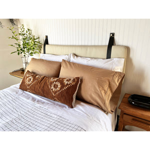 Neutral earth tones bed linen and pillows resting against the sand leather headboard cushion with black straps.