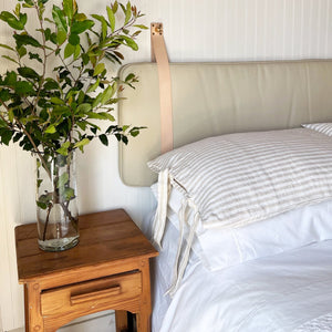 close-up of the wooden side table with clear glass vase holding branches with green leaves. The Sand Leather Headboard Cushion with Straps hanging on against the off-white wall and bed made with fresh linens.