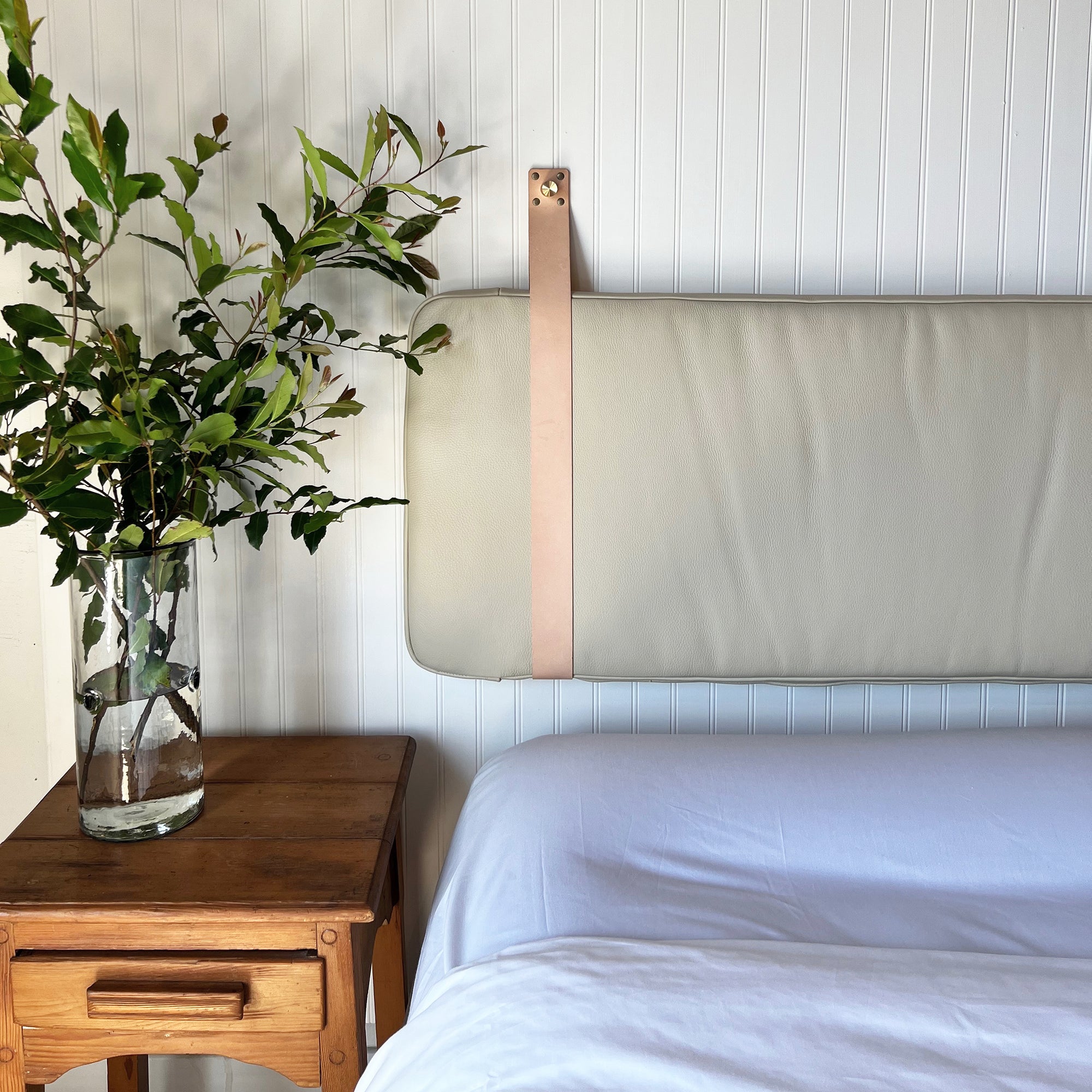 Sand Leather Headboard Cushion with Straps above a bed with no pillows. Wooden side table with a clear glass vase holding branches and leaves.