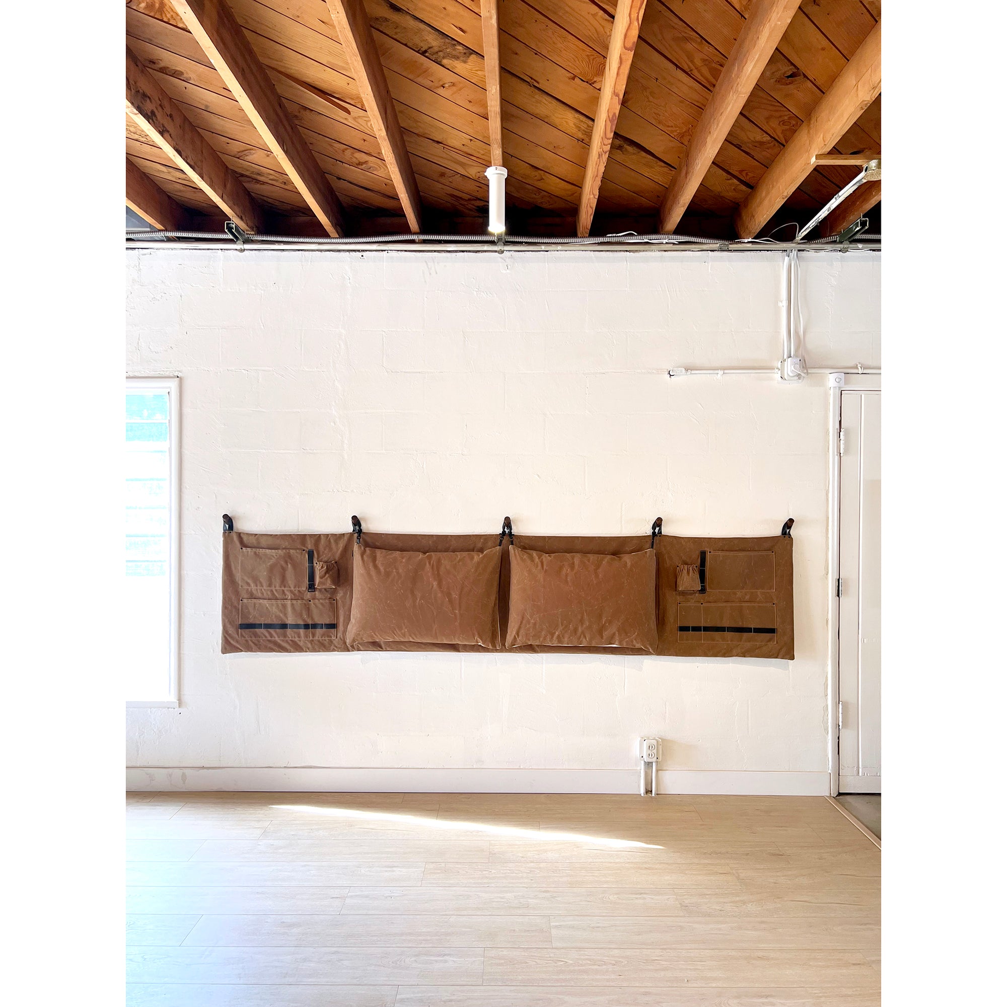 The Scout Headboard in Camel Brown Waxed Cotton Canvas hanging on a white wall in an open studio area. 