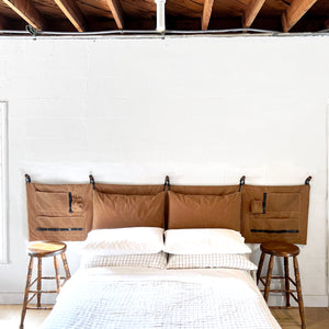 The Scout Headboard - Camel Brown - Waxed Cotton Canvas hanging on a white wall above a queen bed with linen bedding. 