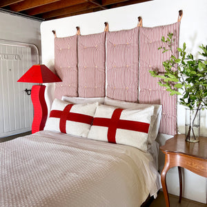 Perspective view of the red and off-white four tuft wall cushion. Natural color leather loops hold the cushions up on the dark wooden pegs on the wall above a bed. 