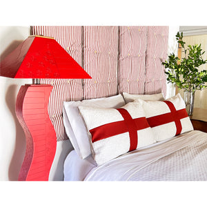 Curvy red  floor lamp next to a made bed with white faux sheepskin and red accent pillows. Tuft wall cushion system is displayed behind the bed.