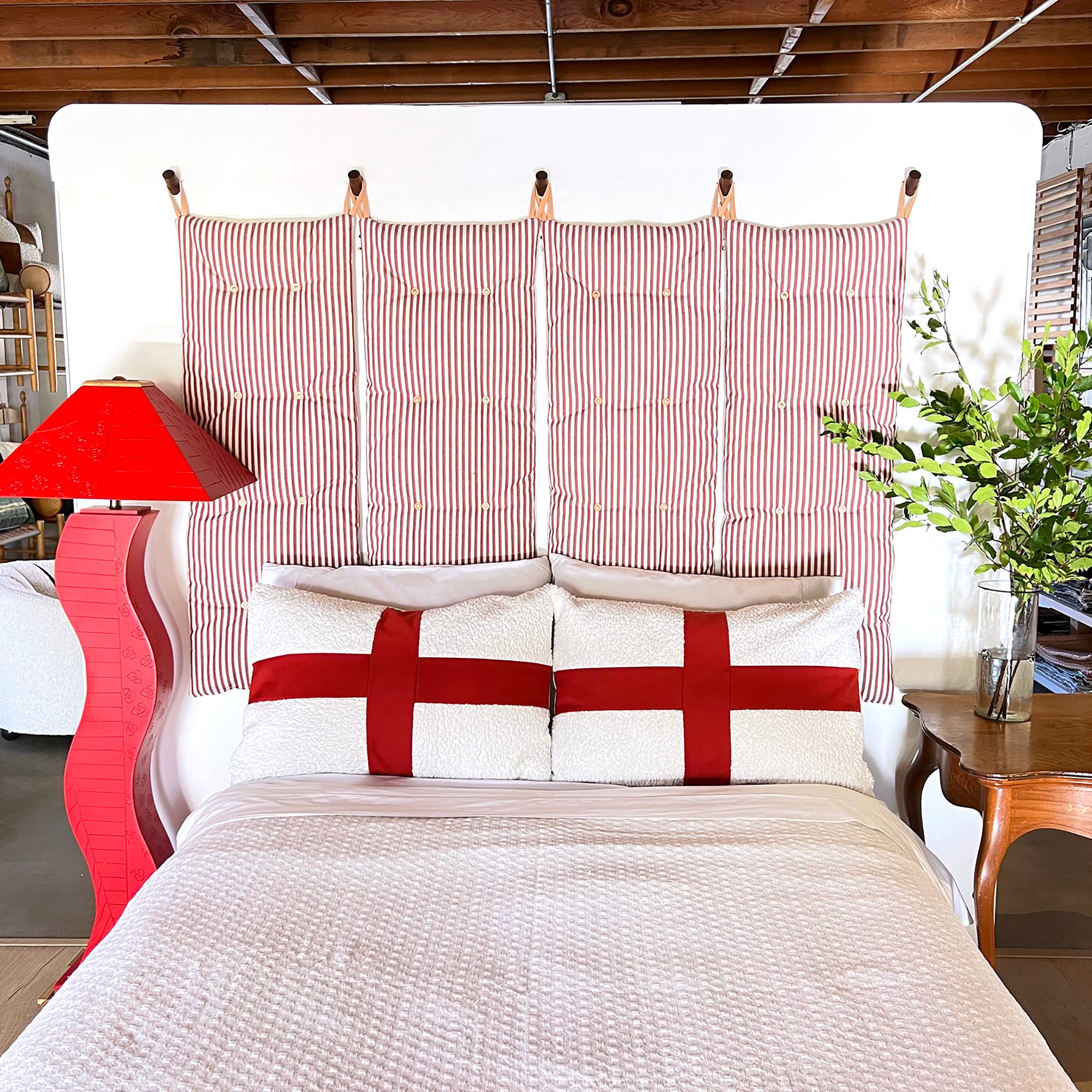 Four red and off-white stripe tuft wall cushions hanging above a queen bed. Red abstract curvy floor lamp to the left and side table with greenery to the right of the bed. 